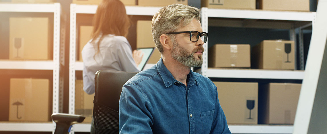 Person working at a computer in a warehouse with shelves of packaged goods, representing ecommerce fulfilment and business growth.